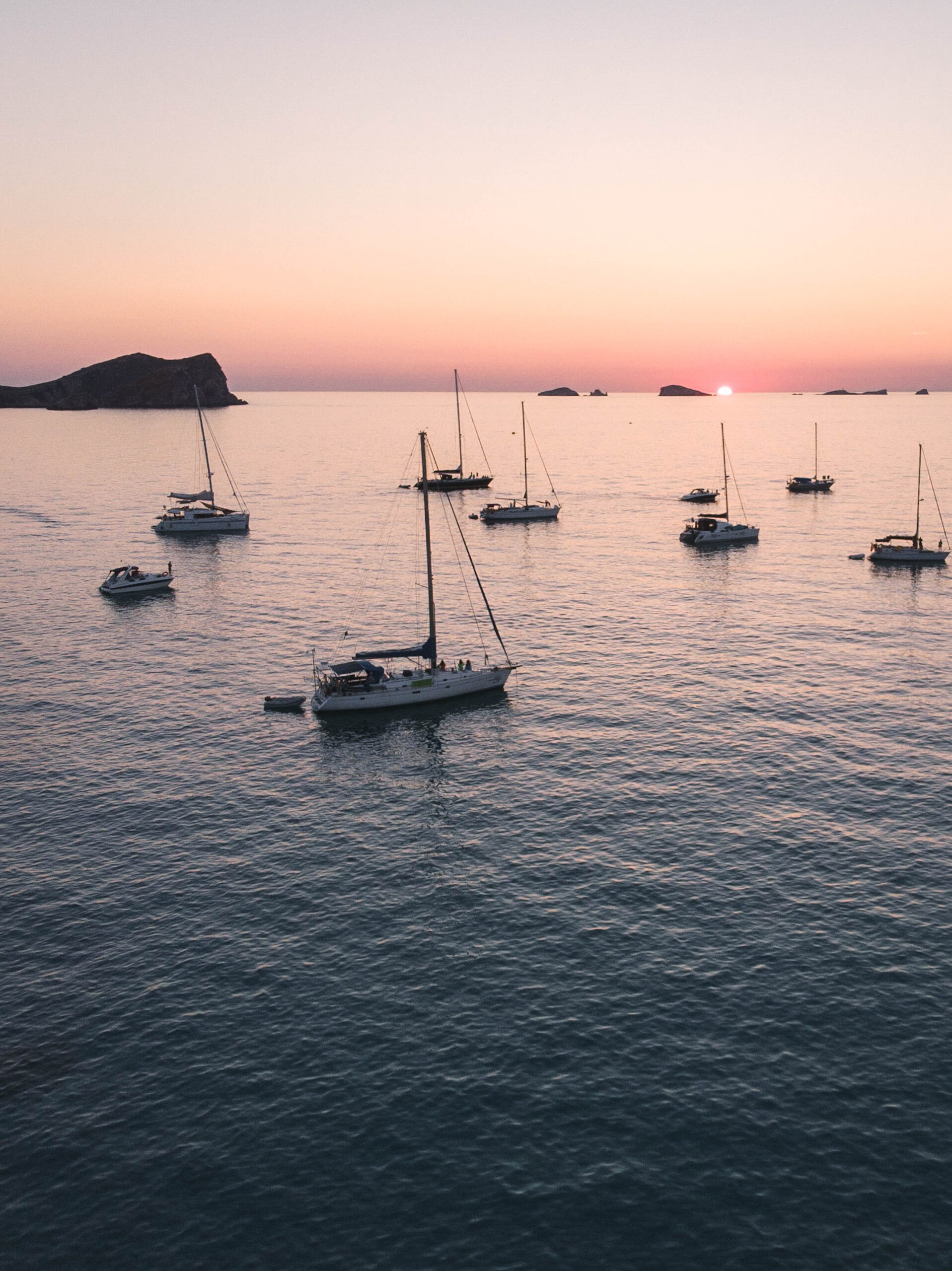 Sailboats anchored at sea during a soft pink sunset off the coast of Ibiza.