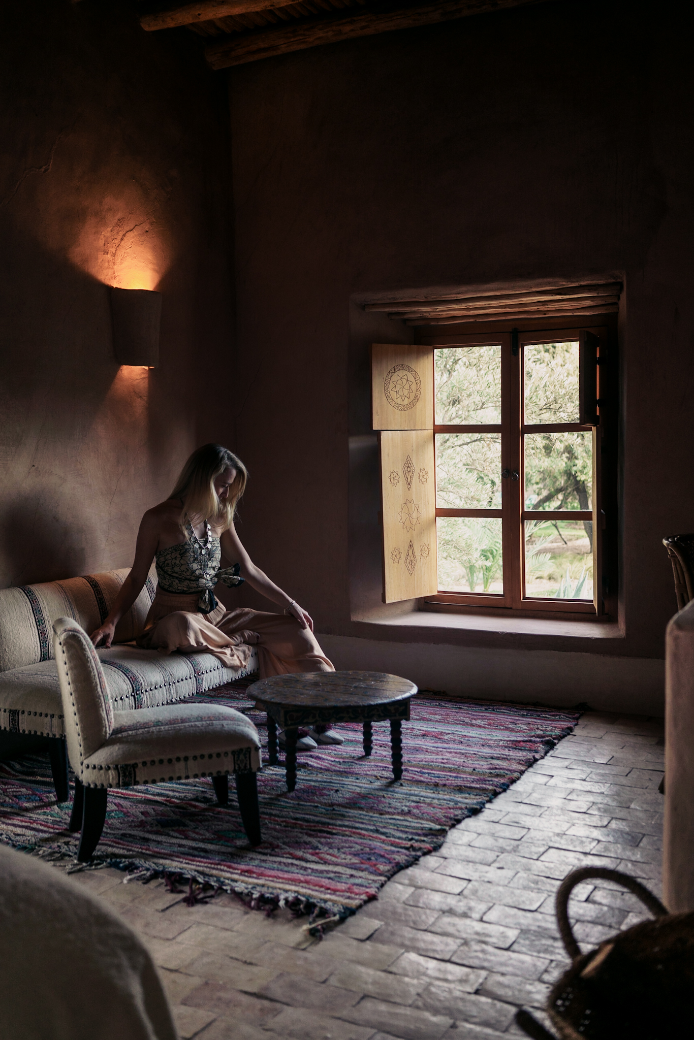 A woman seated on a low sofa in a dim, earth-toned room at a Moroccan retreat, lit softly by a window that opens onto trees.